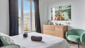 A modern bedroom with a bed, a green chair, a wooden dresser, and a large window with curtains. - Le Léon Gambetta, appartement avec terrasse vue mer (Dieppe)