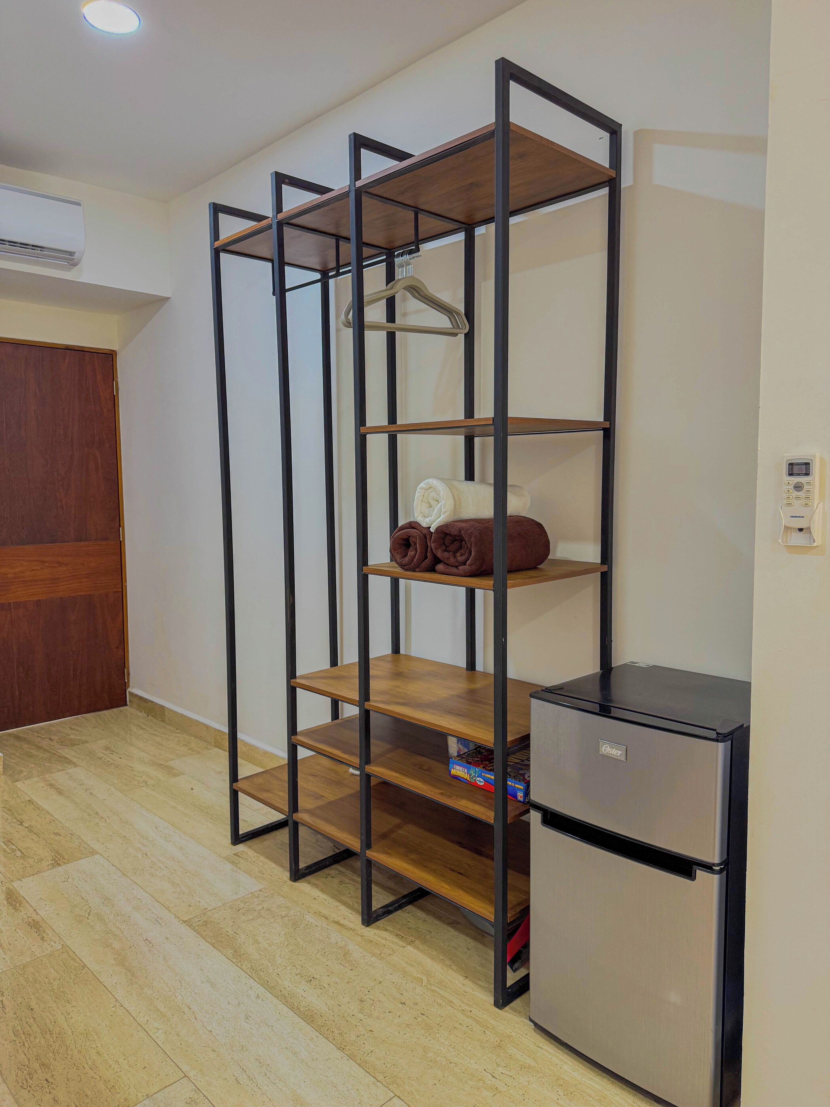 A compact room with a metal shelving unit, a small refrigerator, and a wooden wardrobe.