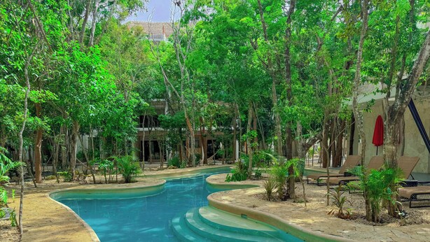 A swimming pool with a stone border, surrounded by trees and a building in the background. - Increible Studio Con Jacuzzi Naia C19 (Tulum)