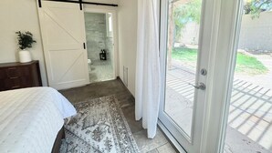 Un dormitorio con cama, una puerta corredera al baño y vistas de un jardín a través de una puerta de vidrio.
