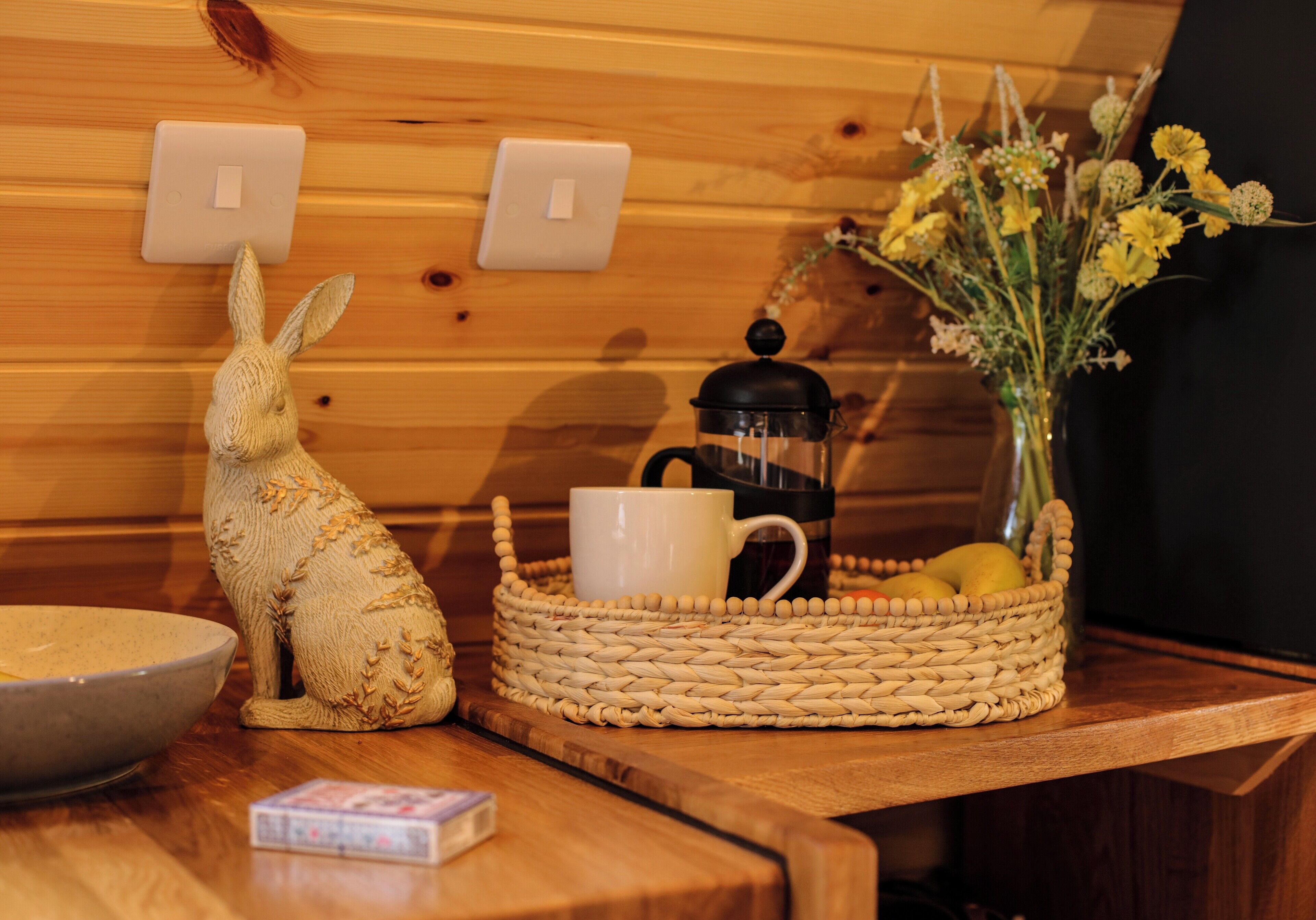 Una mesa de madera con una cesta tejida, una figurita de conejo de cerámica, una taza blanca, una cafetera y un jarrón con flores amarillas.