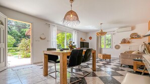 Ein Essbereich mit einem Holztisch und Stühlen, einer Couch und einem kleinen Schrank. Zwei Fenster mit Vorhängen, ein Deckenventilator und eine Pendelleuchte sind vorhanden.