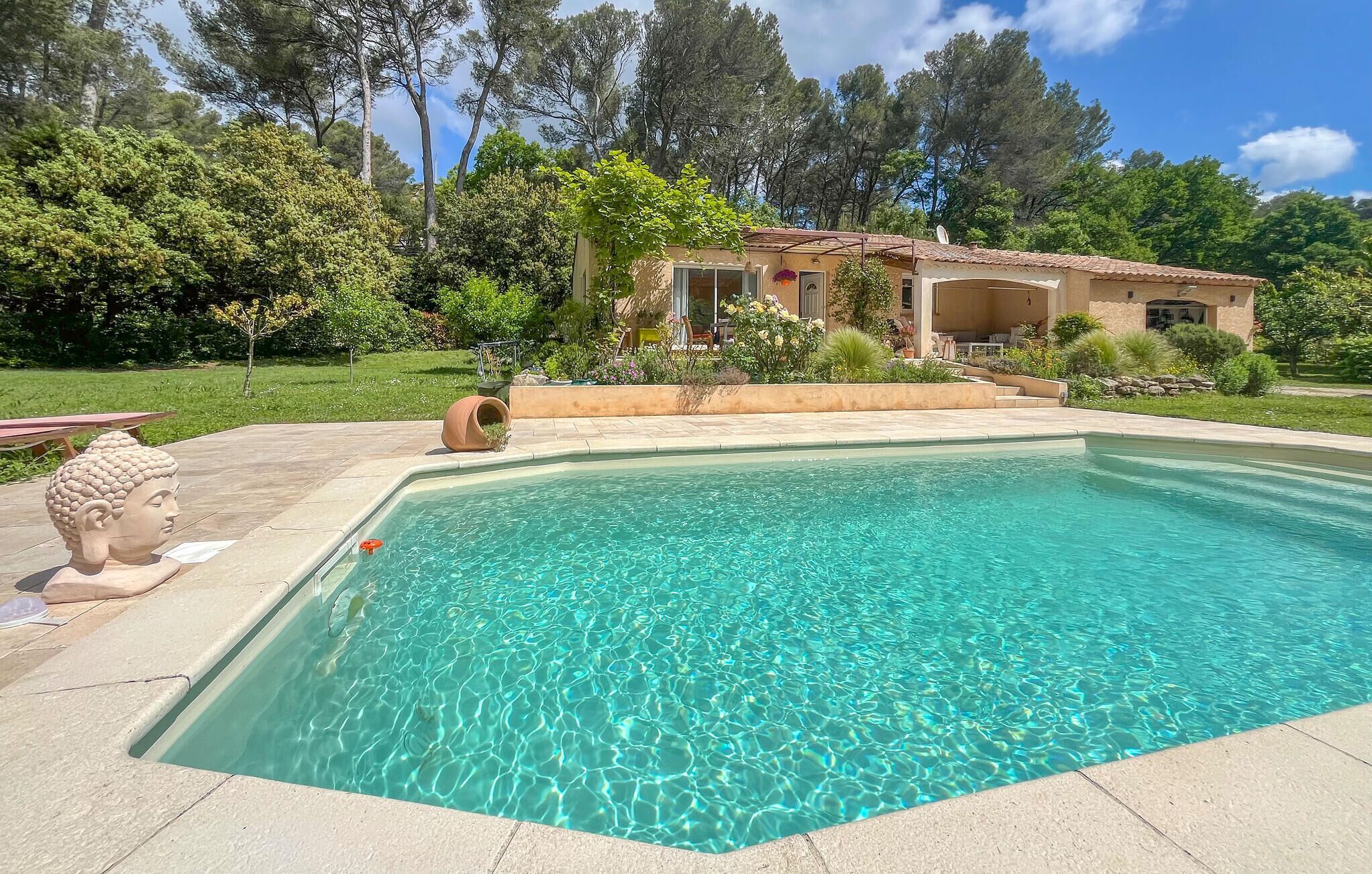 A swimming pool with clear water, surrounded by a garden and a house with a tiled roof.