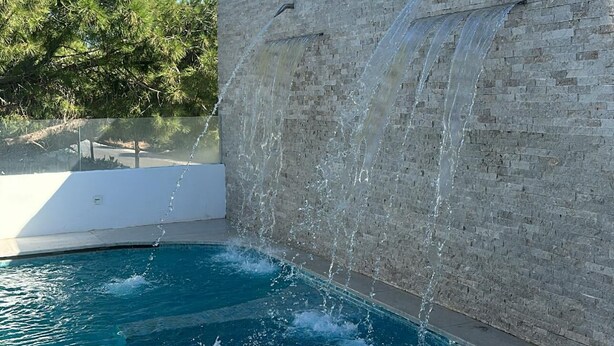 Una piscina con cascata, circondata da un muro in pietra e alberi.