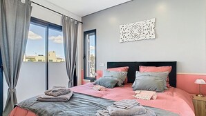 Een moderne slaapkamer met een groot bed, beddengoed in grijze en roze tinten, een decoratief kunstwerk aan de muur, en een uitzicht op gebouwen door het raam.