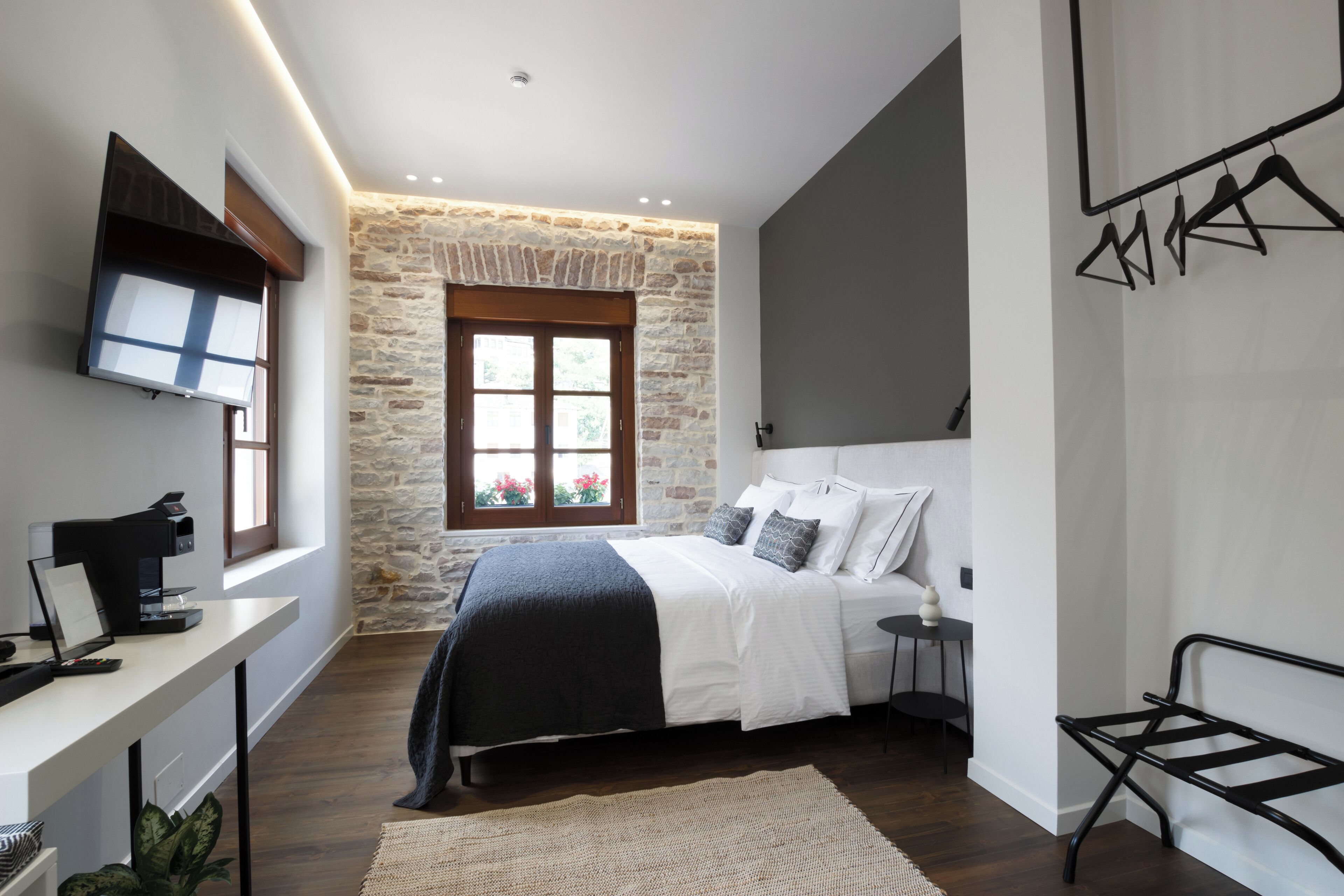 A bedroom with a stone wall, a bed with white bedding, a desk with a computer, and a TV mounted on the wall.