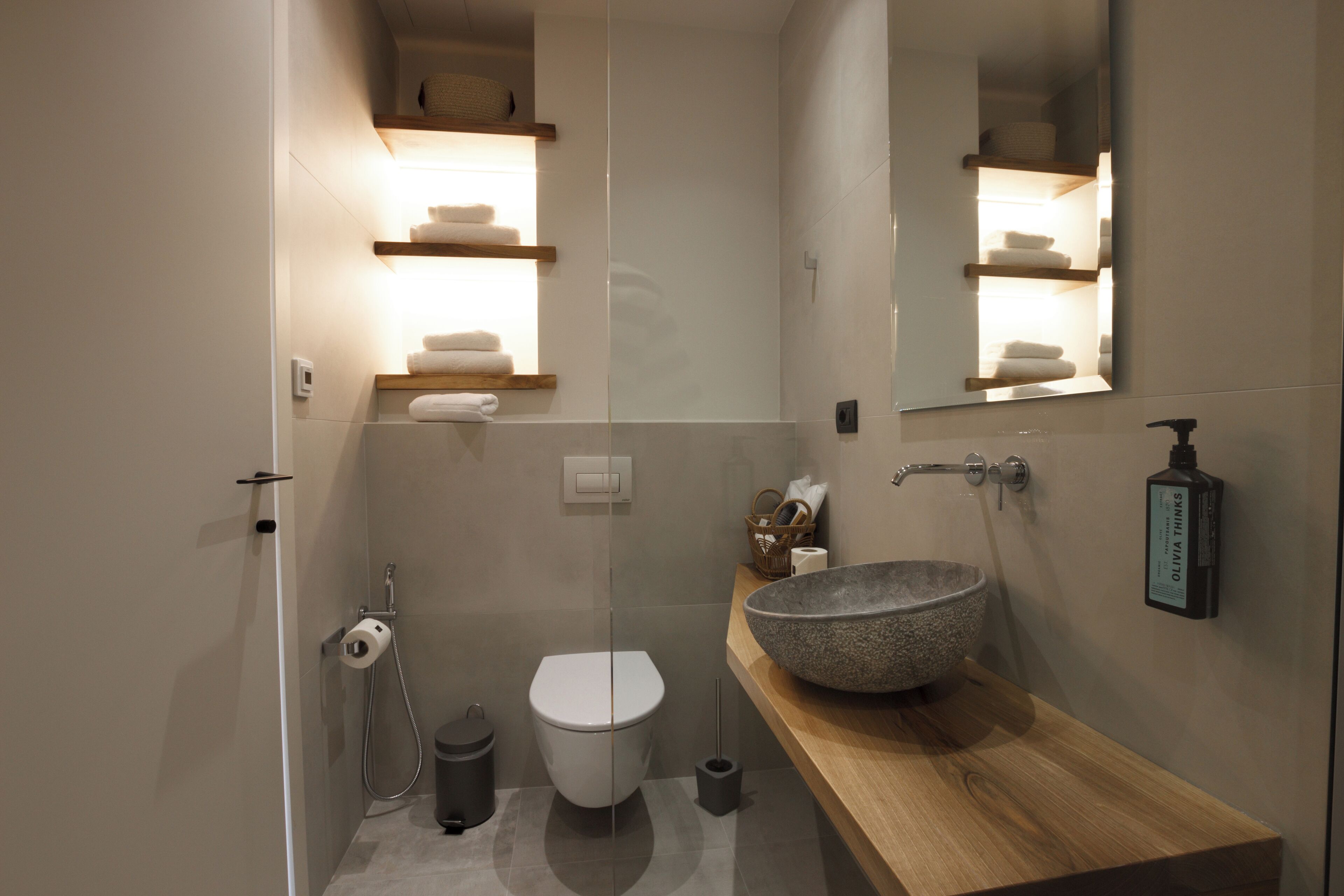 Un baño moderno con un lavamanos de piedra grande, estantes de madera con toallas, un inodoro y un espejo.
