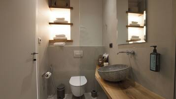 Un baño moderno con un lavamanos de piedra grande, estantes de madera con toallas, un inodoro y un espejo.