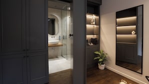 A modern bathroom with a large mirror, a sink, and a shower area. There is a potted plant and a shelf with toiletries. - Stitch&Stone Suites (Gjirokastër)