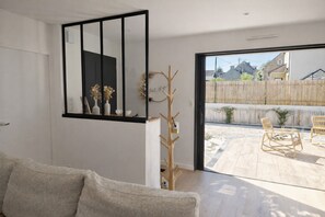 A modern living room with a sofa, a coat rack, and a sliding glass door leading to an outdoor patio with chairs.