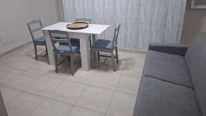 Un comedor con una mesa blanca y cuatro sillas, un sofá gris y piso de baldosas.