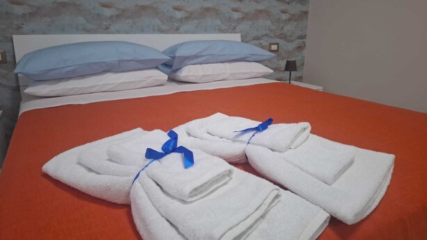 Una cama bien hecha con toallas blancas atadas con cintas azules, una colcha roja y un papel tapiz estampado al fondo.