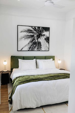 Un lit bien fait, avec une tête de lit verte, une literie blanche et une œuvre d’art représentant un palmier au-dessus.