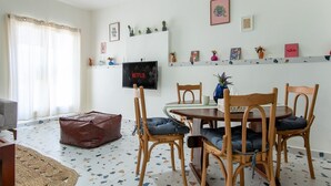 A living room with a round wooden table, four wooden chairs, a sofa, a television mounted on the wall, and a window with curtains.