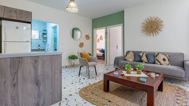 A modern living room with a grey sofa, a coffee table, and a kitchen area with a fridge and sink.