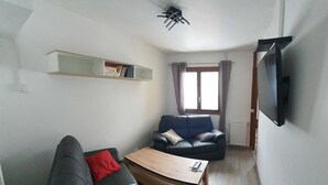 A compact living space with a sofa, a coffee table, a bookshelf, a window with curtains, and a TV mounted on the wall.