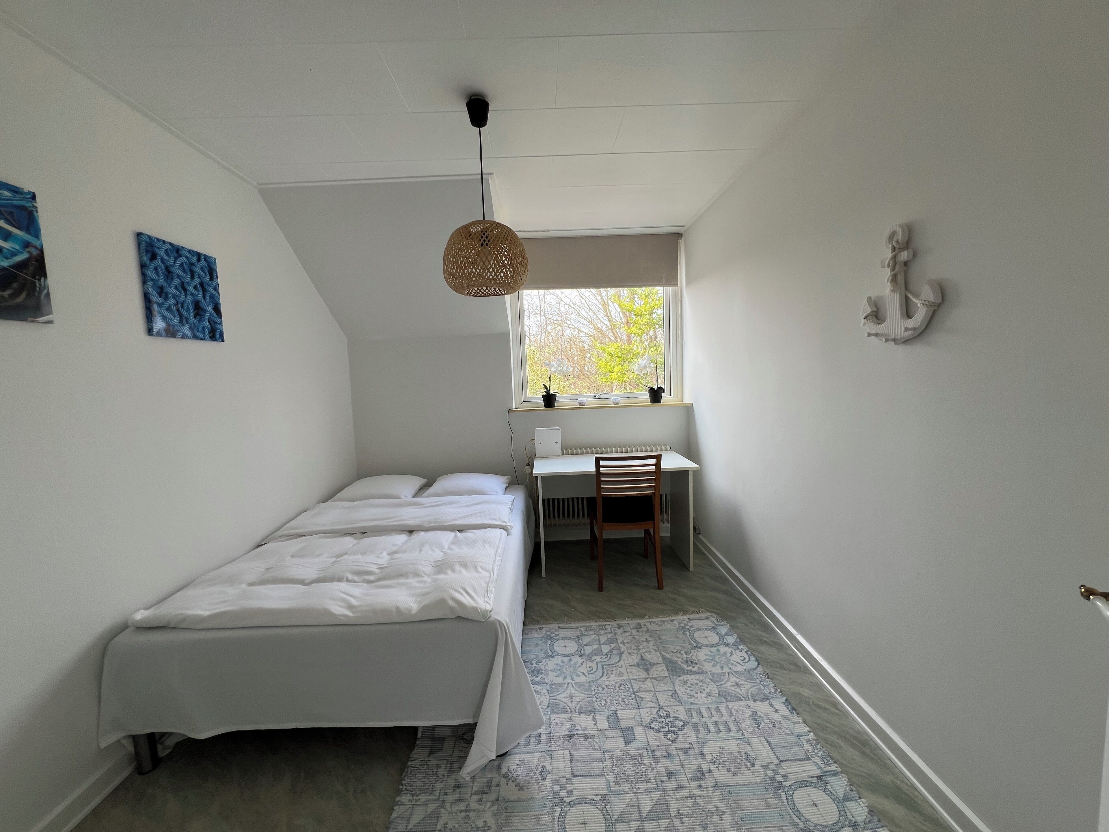 A bedroom with a bed, a desk, and a chair. There is a window with a view of trees and a hanging light fixture.