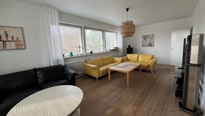 A modern living room with a yellow sofa, a black leather couch, and a white coffee table. There is a large window with blinds, a hanging light fixture, and a refrigerator. - Cozy holiday home (Nakskov)