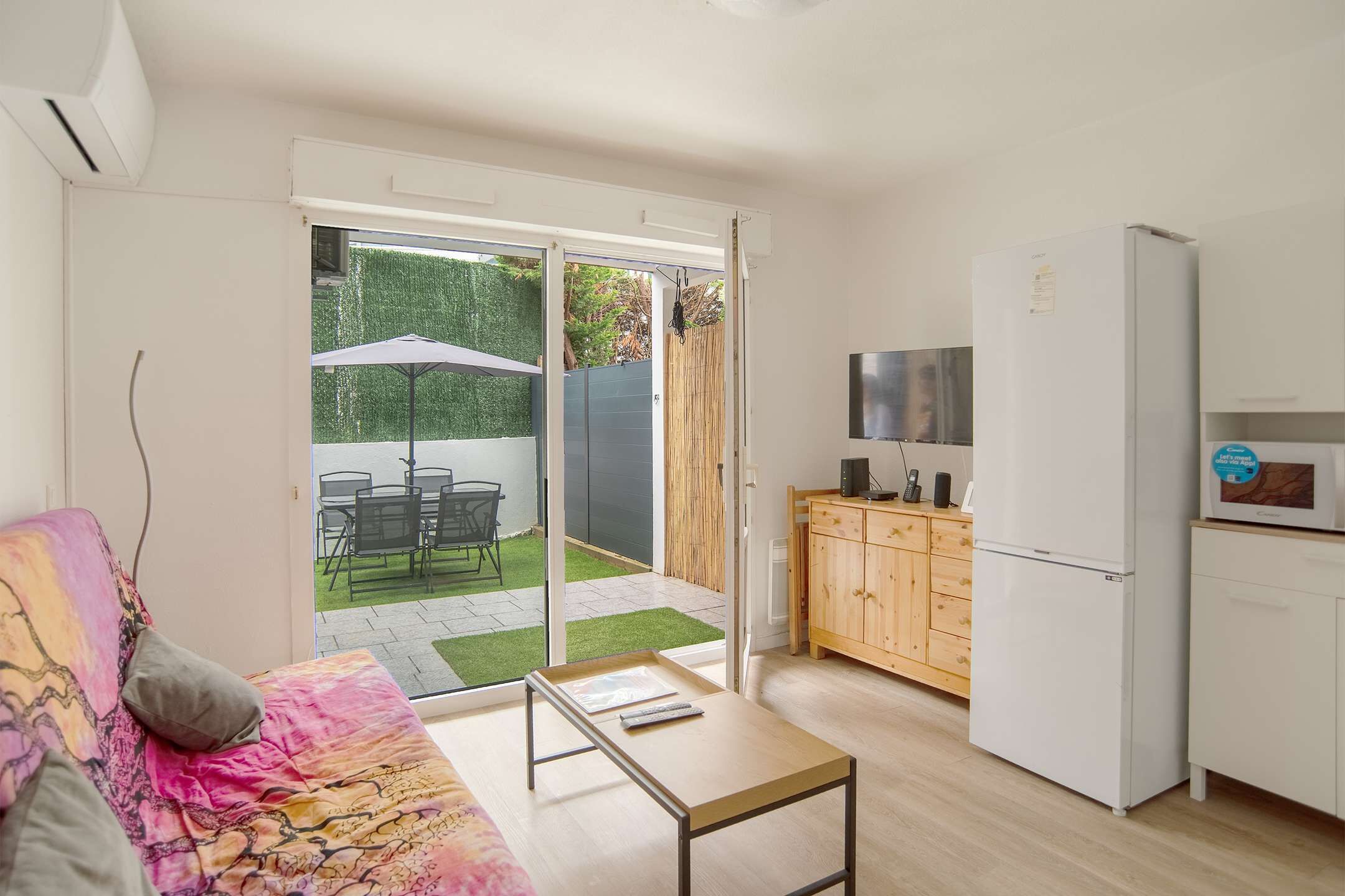 A modern apartment with a sofa, a small table, a kitchen area with a refrigerator, and a sliding glass door leading to a patio with a table and chairs.