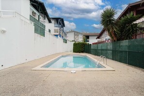 Piscina com escada de metal, cercada por um muro branco e uma cerca verde.