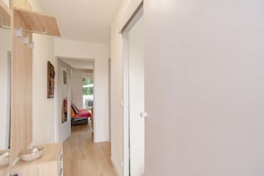 Um corredor com piso de madeira, uma porta branca e vista para um quarto com uma cadeira vermelha e uma mesa.