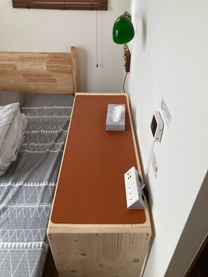 Une chambre à coucher avec une tête de lit en bois, un lit recouvert d’une couverture à motifs, une table de chevet marron avec une boîte de mouchoirs, une lampe de bureau verte et une prise électrique.