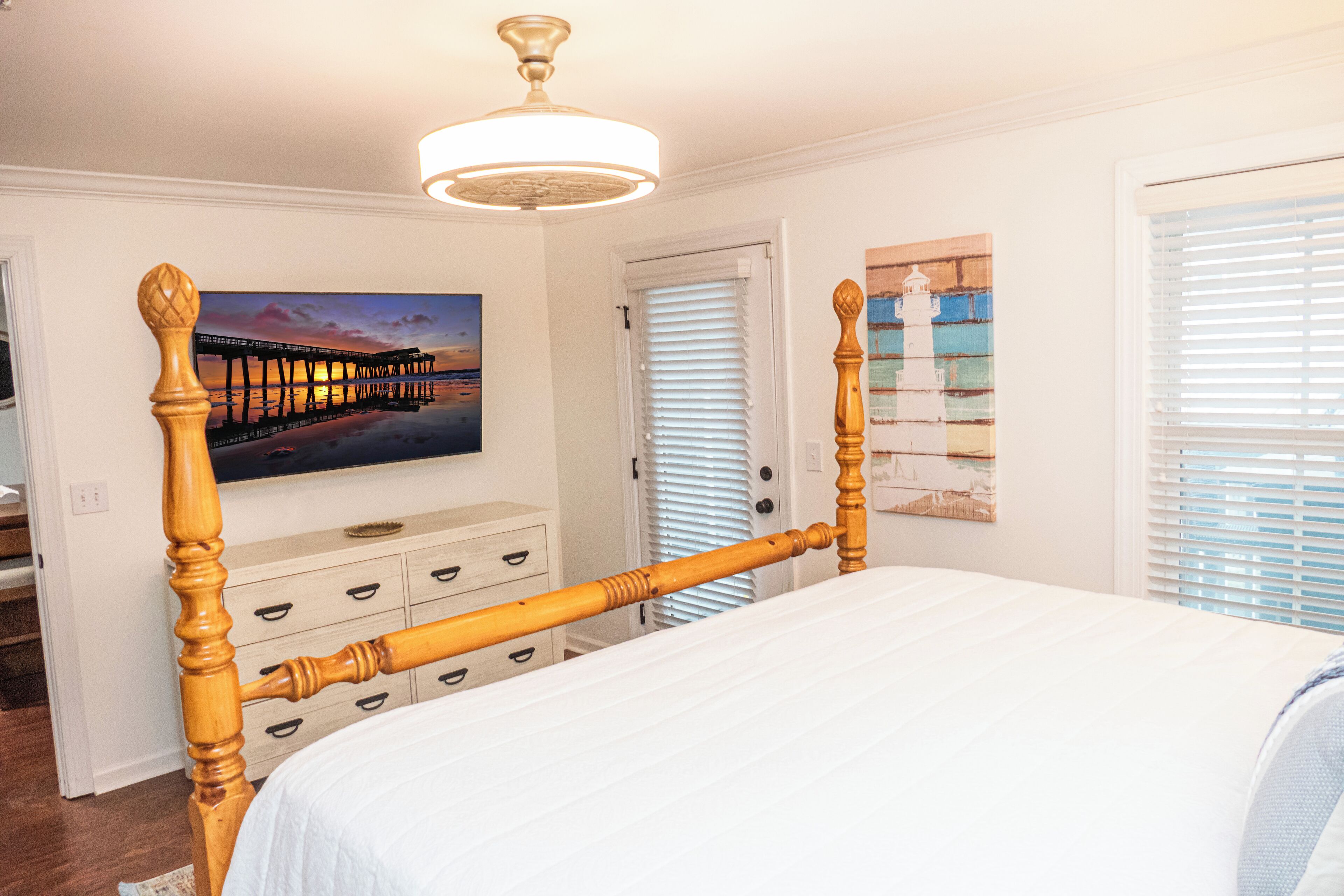 A bedroom with a wooden four-poster bed, a ceiling fan, a painting of a pier, and a door with blinds.