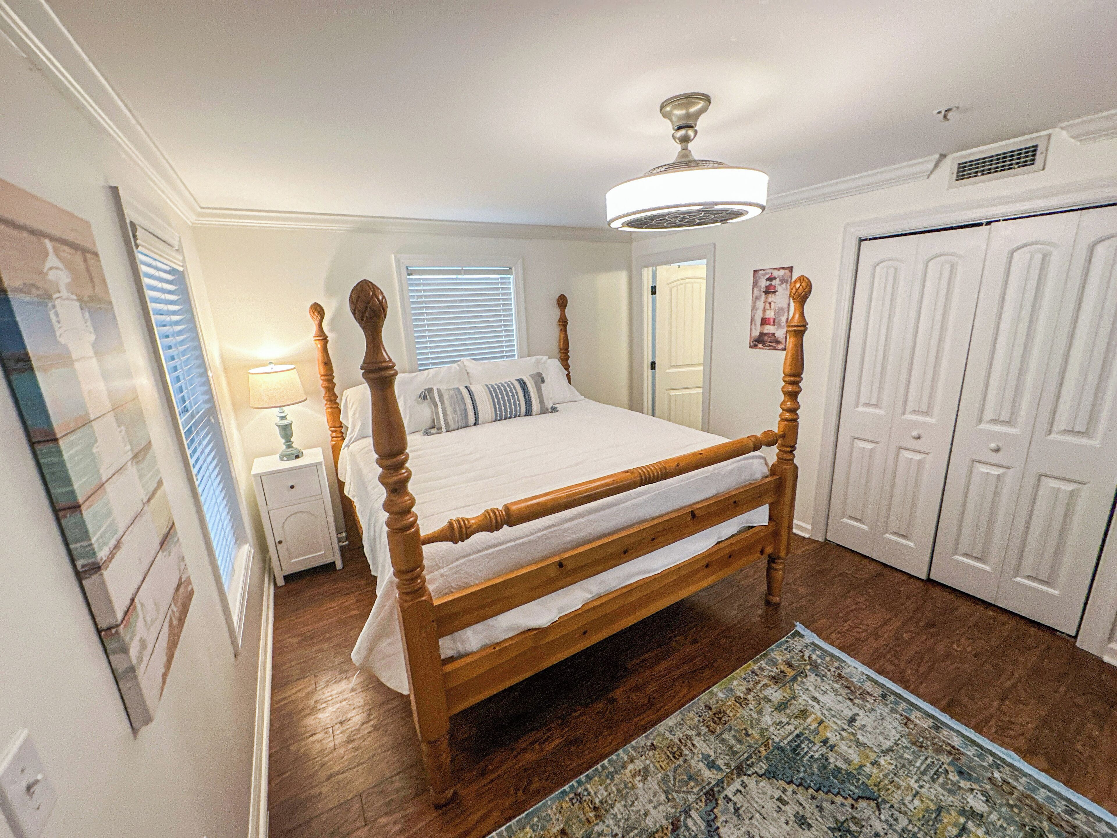 Une chambre à coucher avec un lit à baldaquin en bois, un ventilateur de plafond, une table de chevet avec une lampe et une porte donnant sur une autre pièce.
