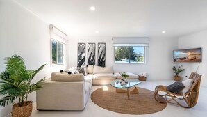 A modern living room with a sofa, coffee table, and a wicker chair. There are large windows and a TV mounted on the wall.