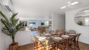 A modern kitchen with a central island, dining area with glass tables and wicker chairs, and a large potted plant.