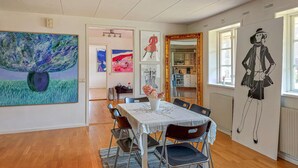 A dining area with a table set for four, surrounded by chairs. There are artworks on the walls, a window with a view, and a radiator.