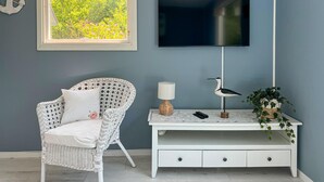 A room with a white wicker chair, a white console table with a lamp and a bird sculpture, a mounted TV, and a window with a view of greenery. - Awesome home in Sturkö with sauna (Sturkö)