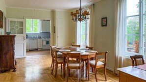 A dining room with a wooden table and chairs, a chandelier, and a view of the garden through large windows.