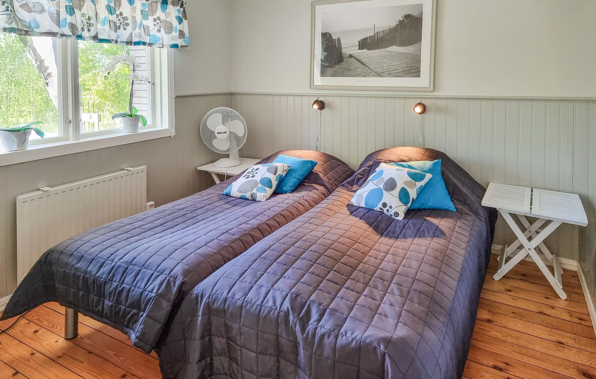 Une chambre avec deux lits, une fenêtre avec un rideau, un radiateur, un ventilateur, une table et un tableau encadré au mur.