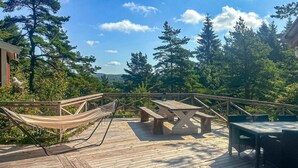A wooden deck with a hammock, picnic table, and chairs, surrounded by trees and a clear sky.