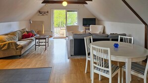 En hyggelig opholdsstue med sofa, spisebord med stole, fjernsyn og udsigt til naturen gennem et stort vindue.