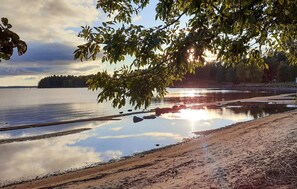 Un lac paisible aux rives sablonneuses, bordé d’arbres et s’étendant jusqu’à une côte lointaine.