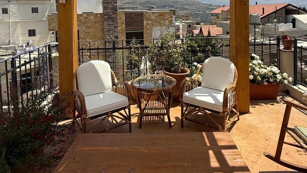 A wooden pergola with two white cushioned chairs and a small round table, overlooking a cityscape with mountains in the distance. - A century of stories, now yours to experience. (Ehden)