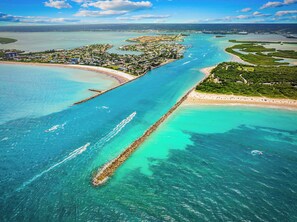 An aerial view of a coastal area with a long pier extending into turquoise waters, a beach, and a residential area with buildings.