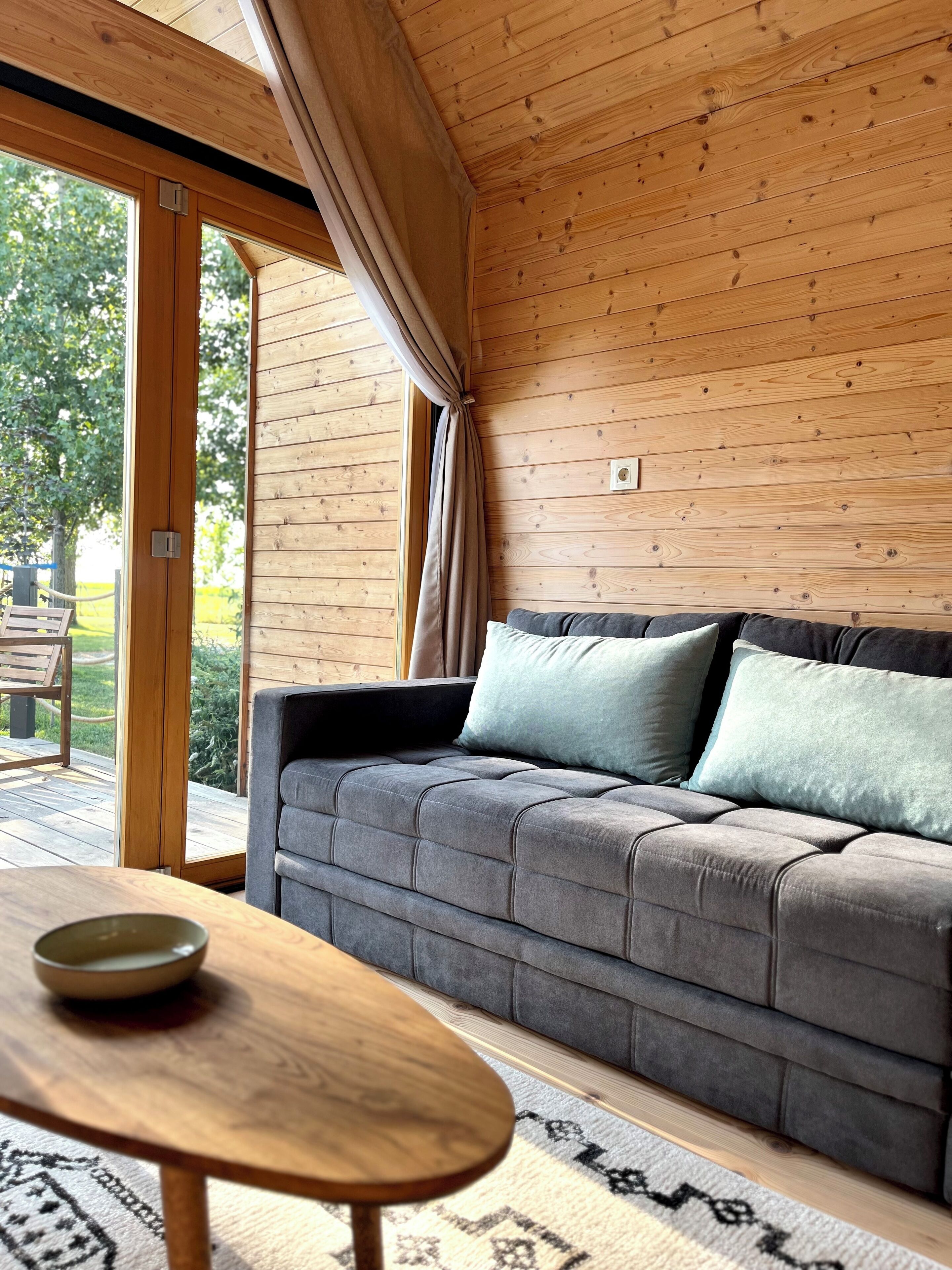 A cozy room with a grey sofa, a wooden table, and a large window with curtains.