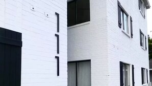 Un bâtiment blanc avec des encadrements de fenêtres et des volets noirs, et deux jardinières noires contenant des arbustes taillés en forme de sphère, le tout placé sur un trottoir.