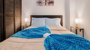A hotel room with a bed, two bedside tables with lamps, a cityscape painting on the wall, and a window with blinds. - Loft La Luz - Old San Juan (San Juan)
