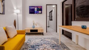 A modern living room with a yellow sofa, a wooden coffee table with a vase, a flat-screen TV mounted on the wall, and a large window with blinds. - Loft La Luz - Old San Juan (San Juan)