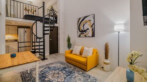 A modern living room with a yellow sofa, a wooden coffee table, a patterned rug, a black spiral staircase, a painting, and a floor lamp. - Loft La Luz - Old San Juan (San Juan)