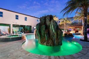 A pool area with a rock centerpiece, lounge chairs, and a building with a green wall and glass doors.
