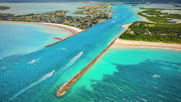 Vista aérea de una zona costera con un muelle largo que se adentra en aguas turquesas, una playa y una zona residencial con edificios.