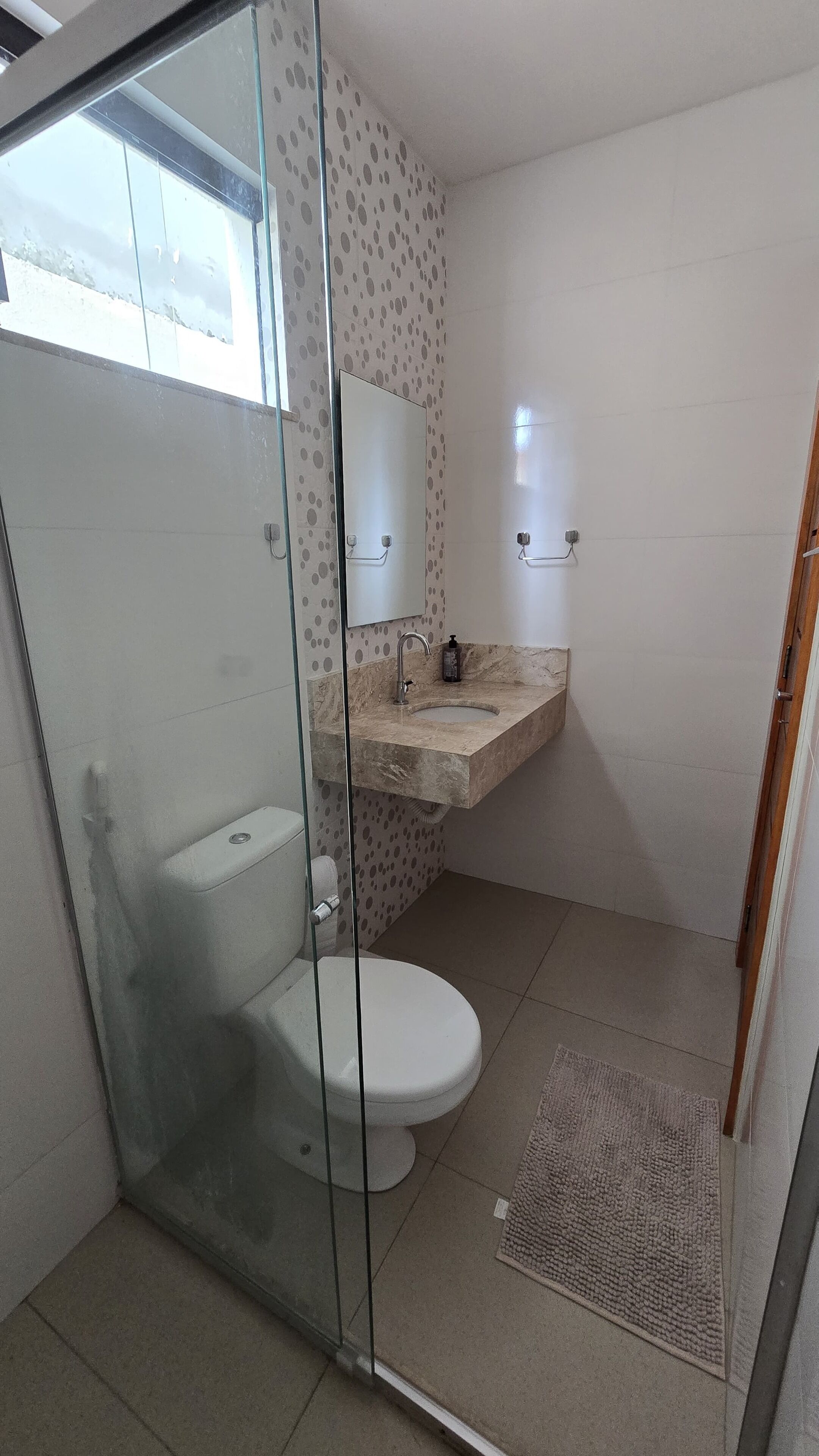 A bathroom with a glass shower enclosure, a toilet, a sink with a marble countertop, and a patterned wall.