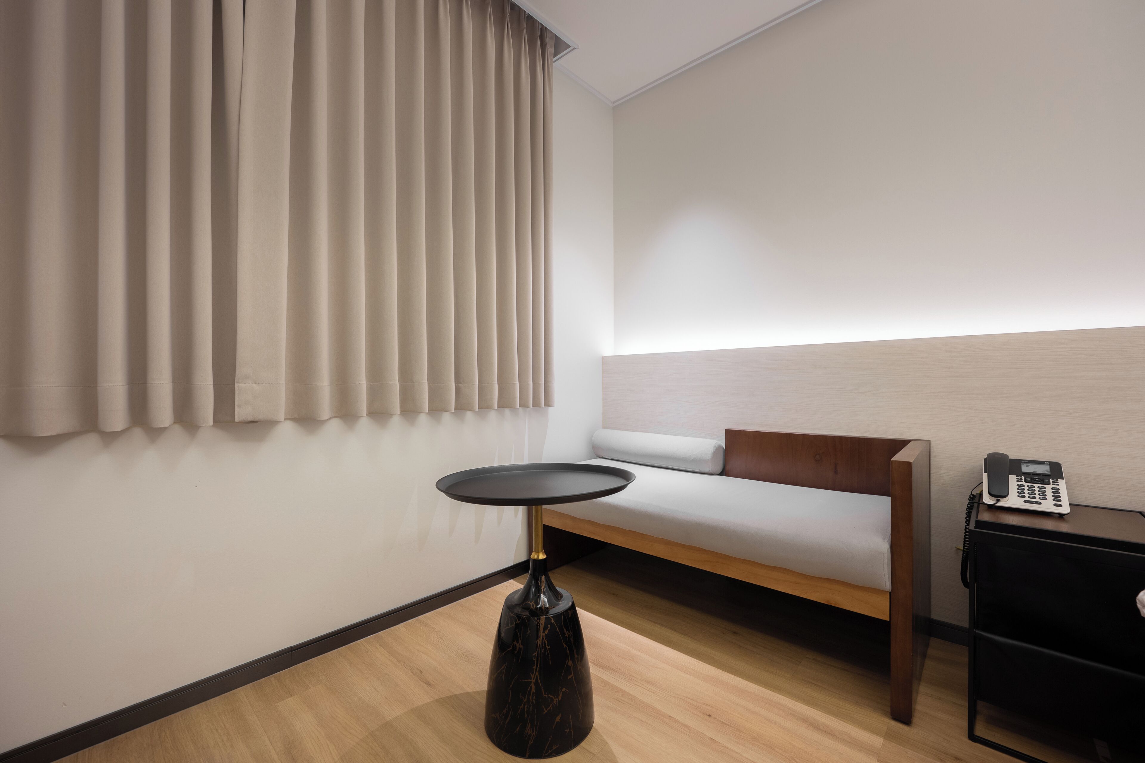 A modern hotel room with a wooden floor, a sofa bed, a small round table, and a telephone on a side table.
