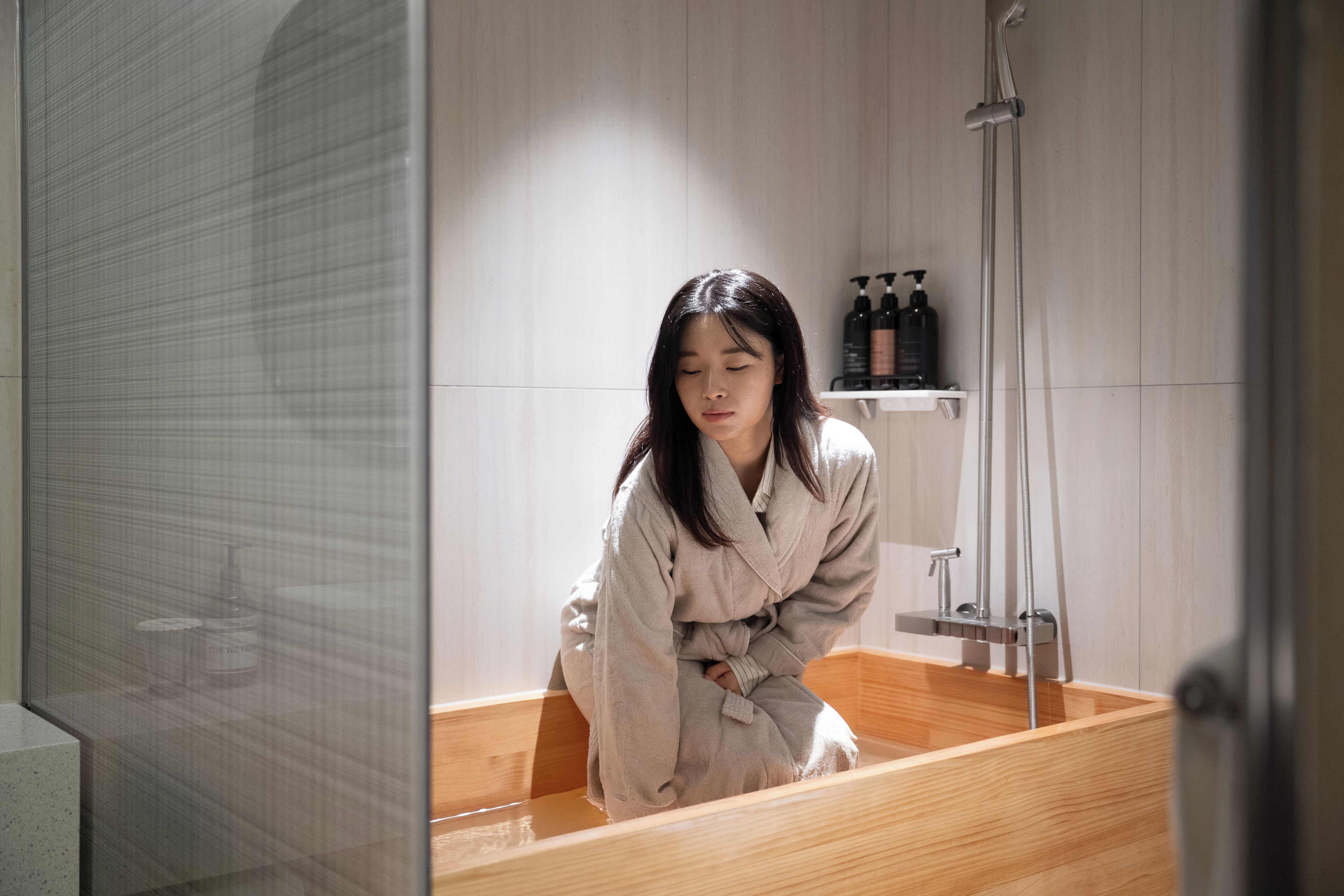 En person i badekåbe sidder i et træbadekar i et moderne badeværelse med bruser og en hylde med toiletartikler.