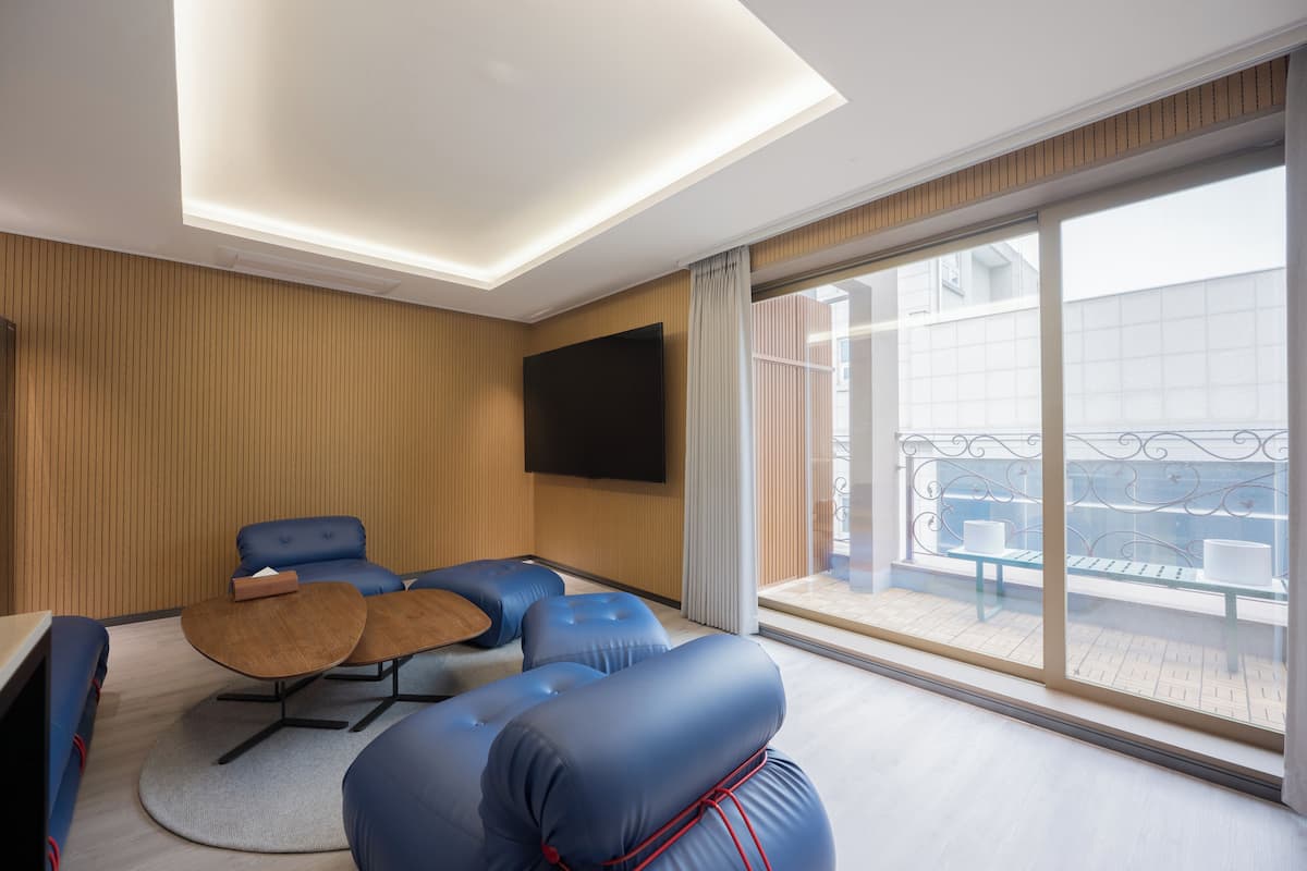 A modern hotel room with a large balcony, a flat-screen TV, and a seating area with blue sofas and a coffee table.